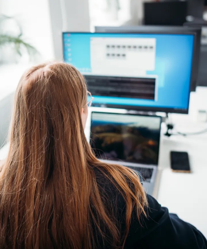 Back view of a woman working on a laptop and desktop computer.