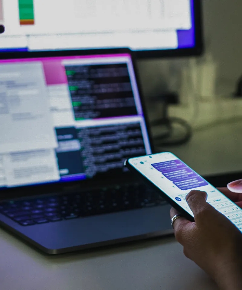 Hands holding a mobile phone in the foreground, and a laptop placed on the desk in the background.