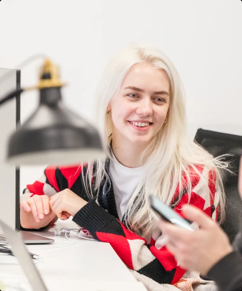 A QA engineer sitting at a desk, chatting with a colleague in the background.