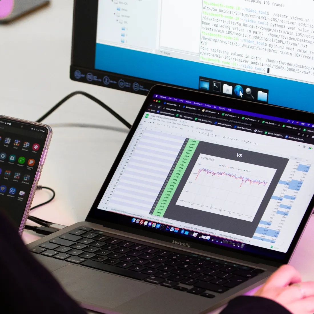 A QA engineer holding a phone and working with a laptop, which is placed on a desk.