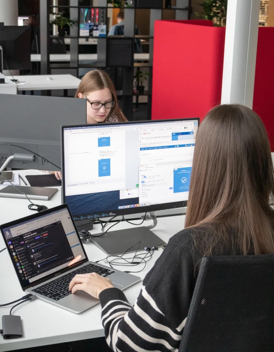 Two QA engineers sitting in front of each other, with monitors and a laptop on the desk.