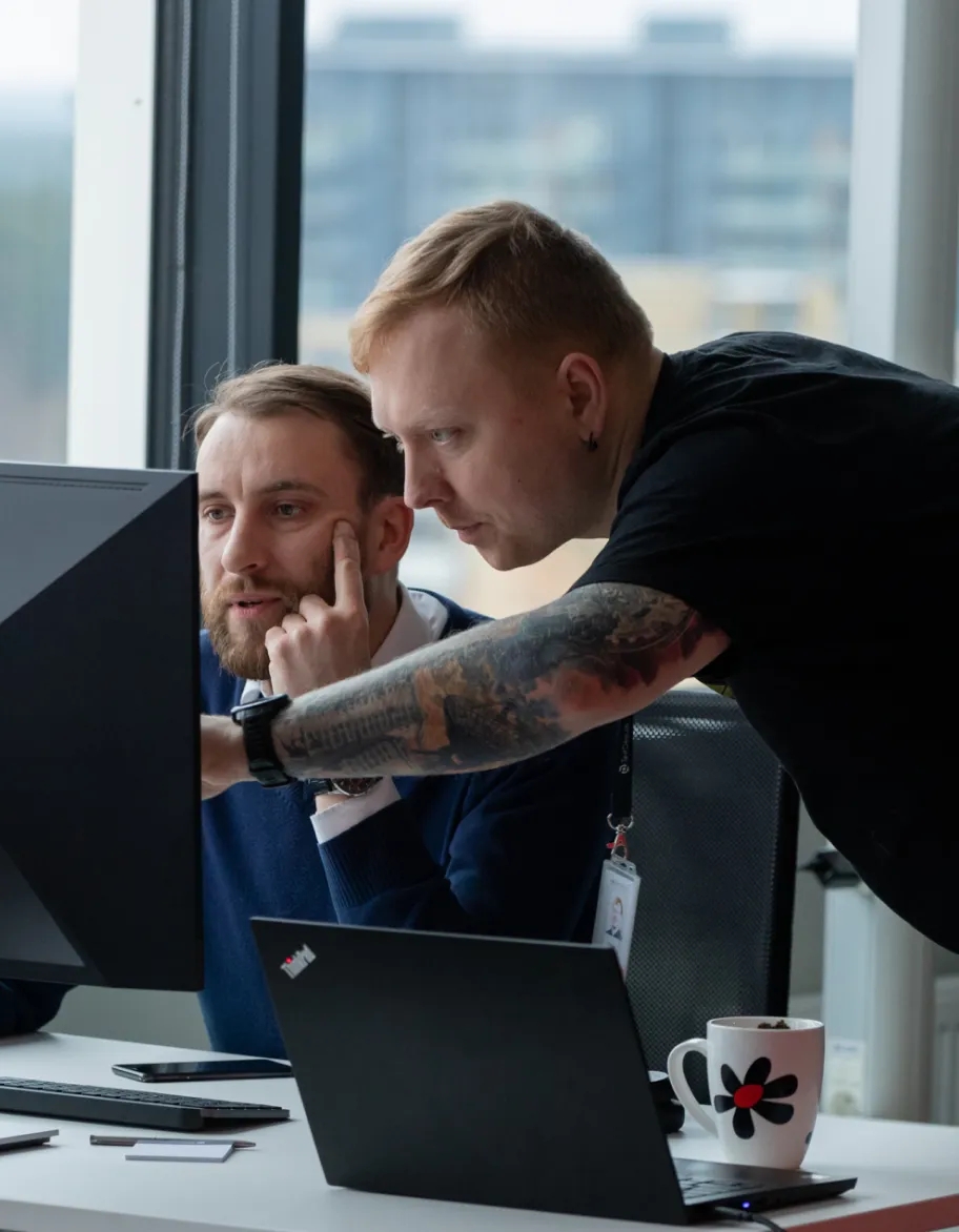 Two QA engineers, where one is pointing at the monitor, but the other is listening.