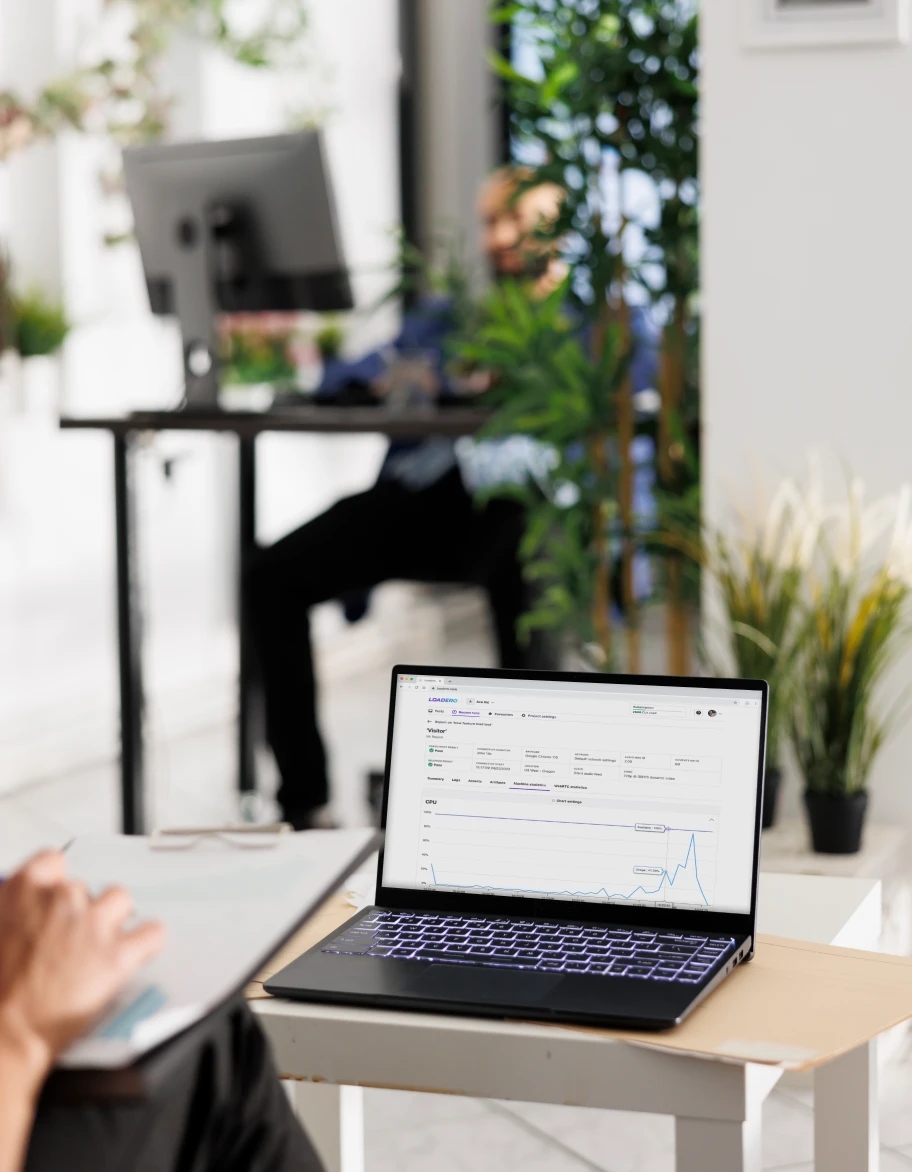 Un espacio de oficina abierto con plantas, con un portátil en un pequeño escritorio mostrando gráficos de datos, y una persona trabajando en el fondo.