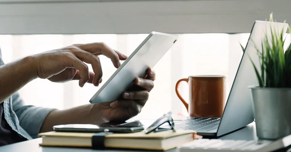 Person using tablet while seated at a desk facing a laptop