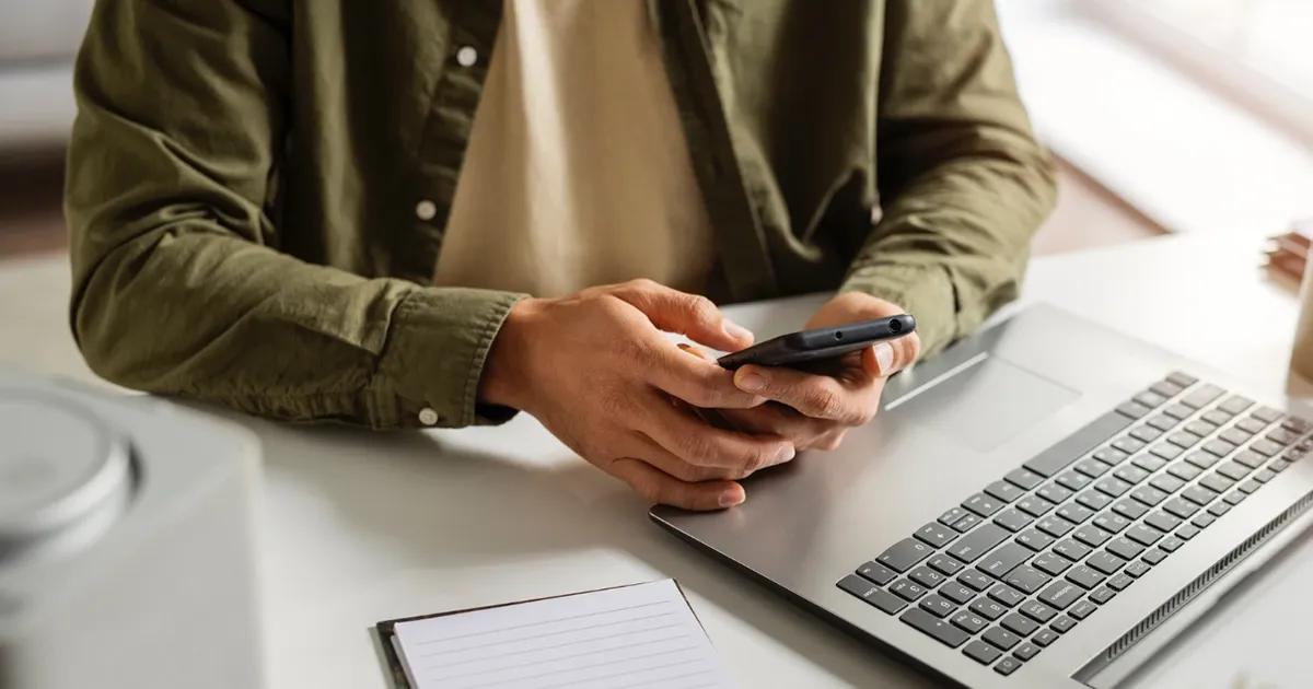 Man sitting at a desk, holding a smartphone, with a laptop open in front of him.