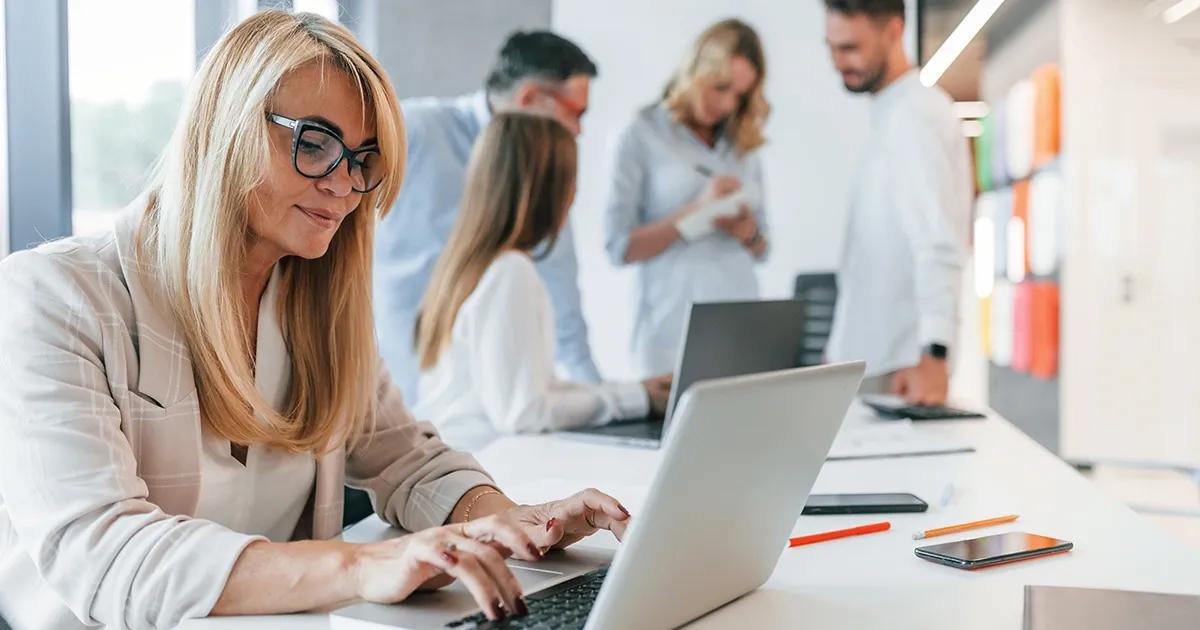 Corporate woman working on laptop at a desk with multiple people in the background