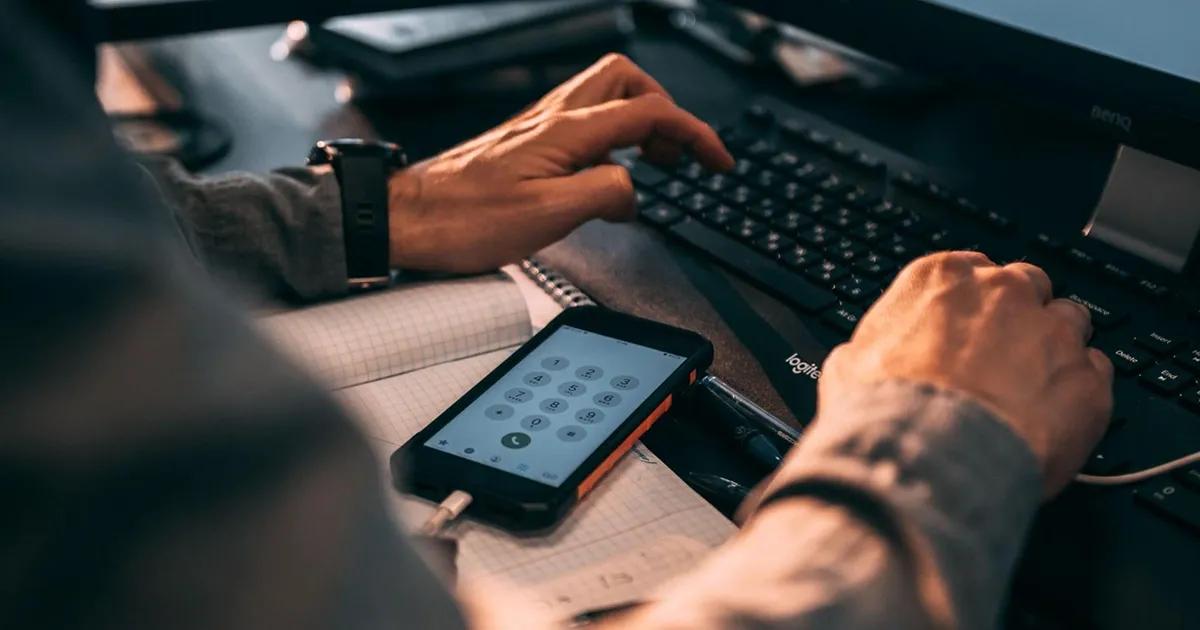 Person typing on a keyboard with a mobile phone in front of them displaying an on-screen keypad