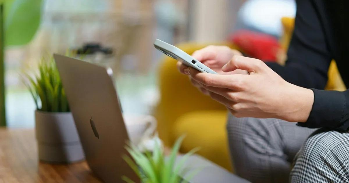 Person using a mobile phone with a laptop placed on a table in front of them