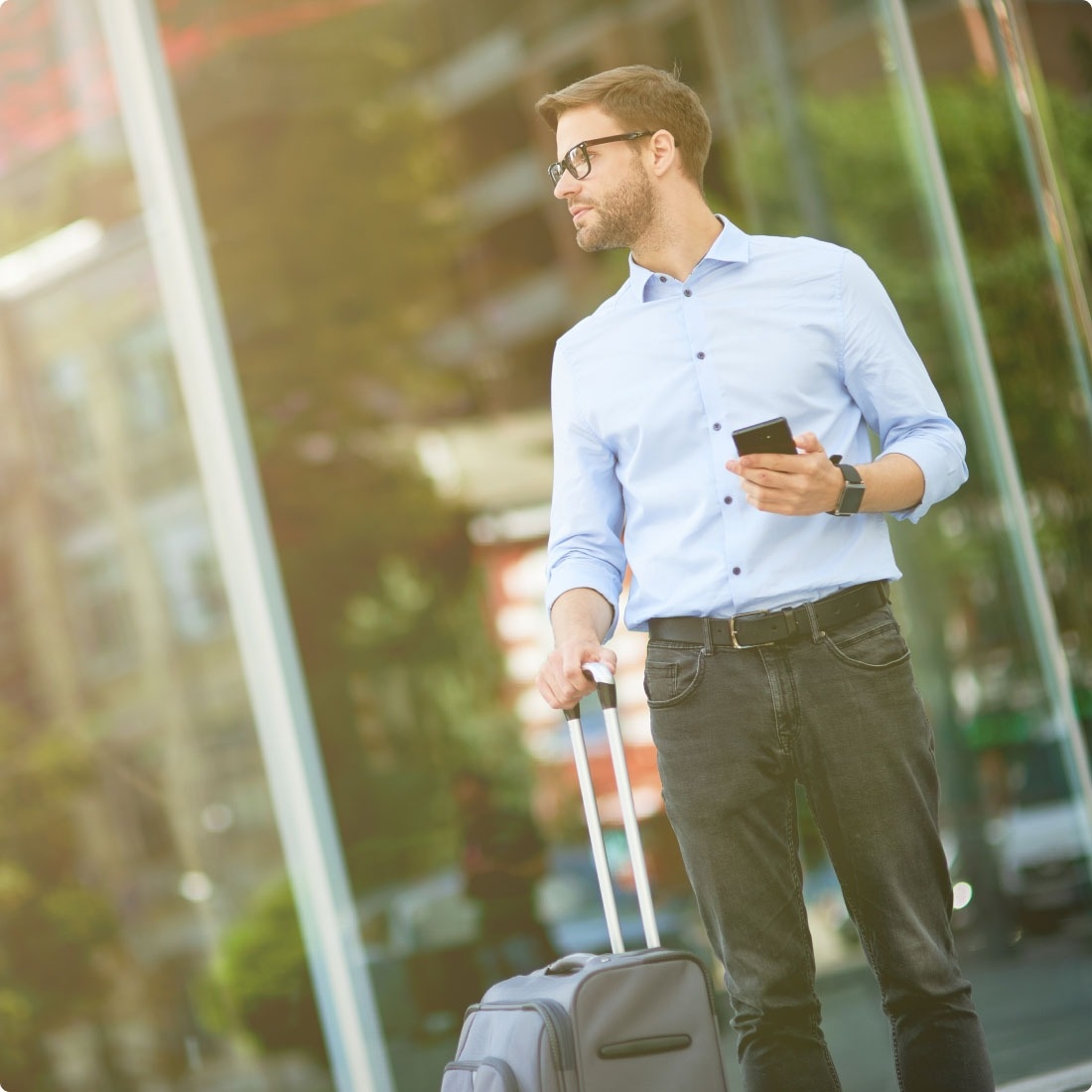 En mand står udenfor med en mobiltelefon og en kuffert i hånden.