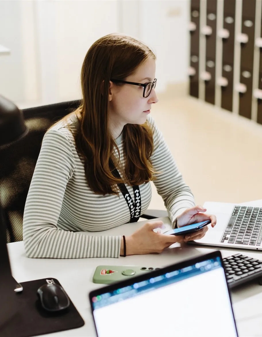 QA inženieris pie galda, kas tur viedtālruni un skatās datora ekrānā.
