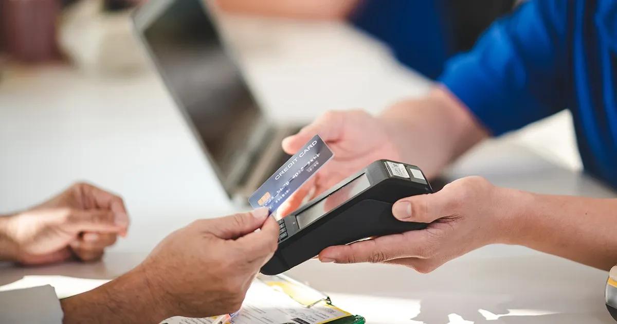 Hand holding a credit card in front of a payment terminal held by another person